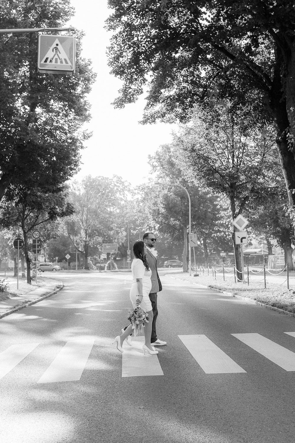 Black and white photo of a couple crossing a zebra crossing with a dog on a tree-lined urban street.