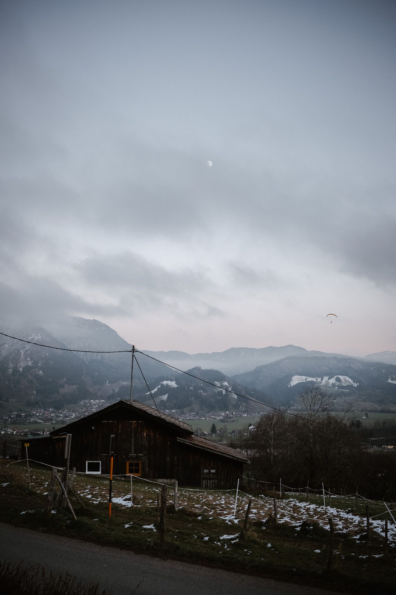 Rustic wooden chalet on snowy hillside with misty mountain valley and paraglider in cloudy winter sky.