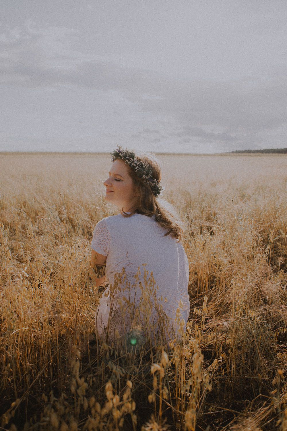 Woman wearing a floral crown standing in a golden wheat field with eyes closed at sunset.