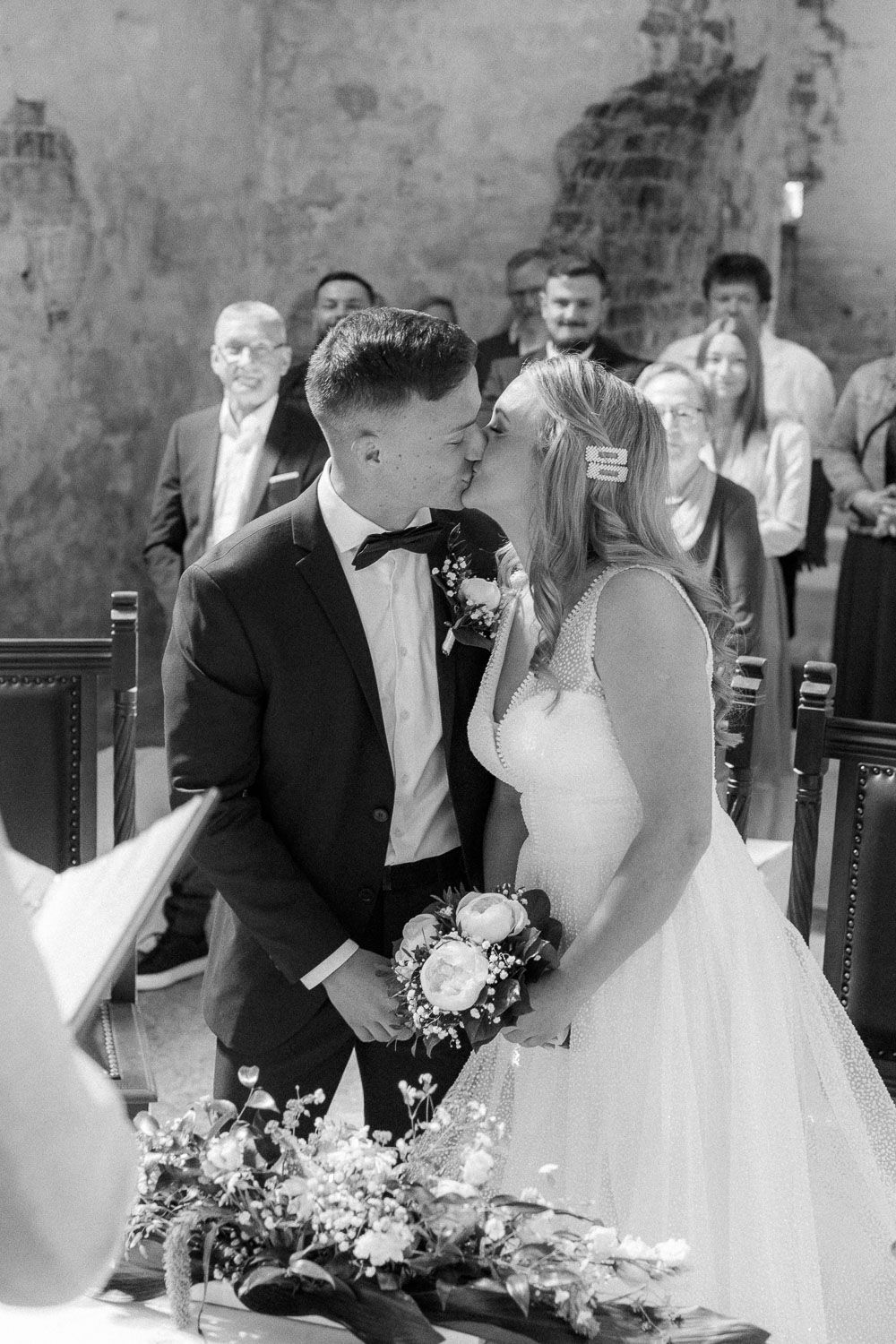 Black and white photo of newlyweds sharing first kiss at altar with guests watching in background.