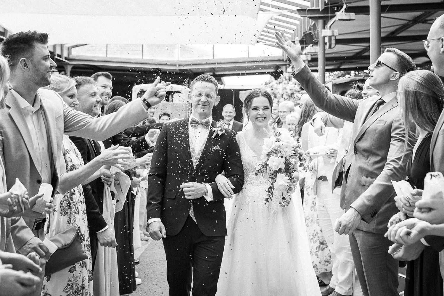 Bride and groom walk through confetti shower surrounded by cheering wedding guests in black and white photo.