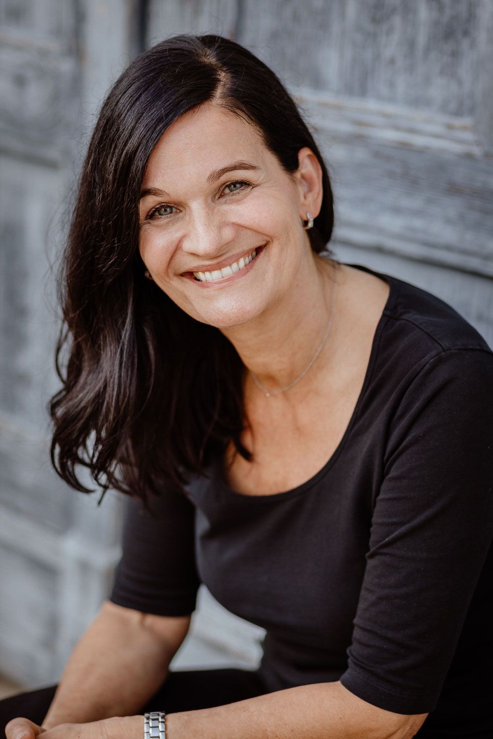Smiling brunette woman in black top seated outdoors against a rustic wooden background.