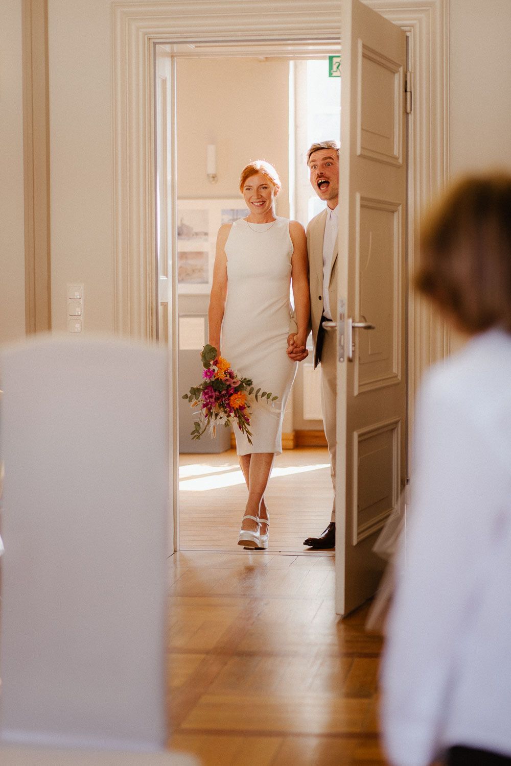 Smiling bride in white sheath dress and groom entering a bright elegant room holding a colorful bouquet.