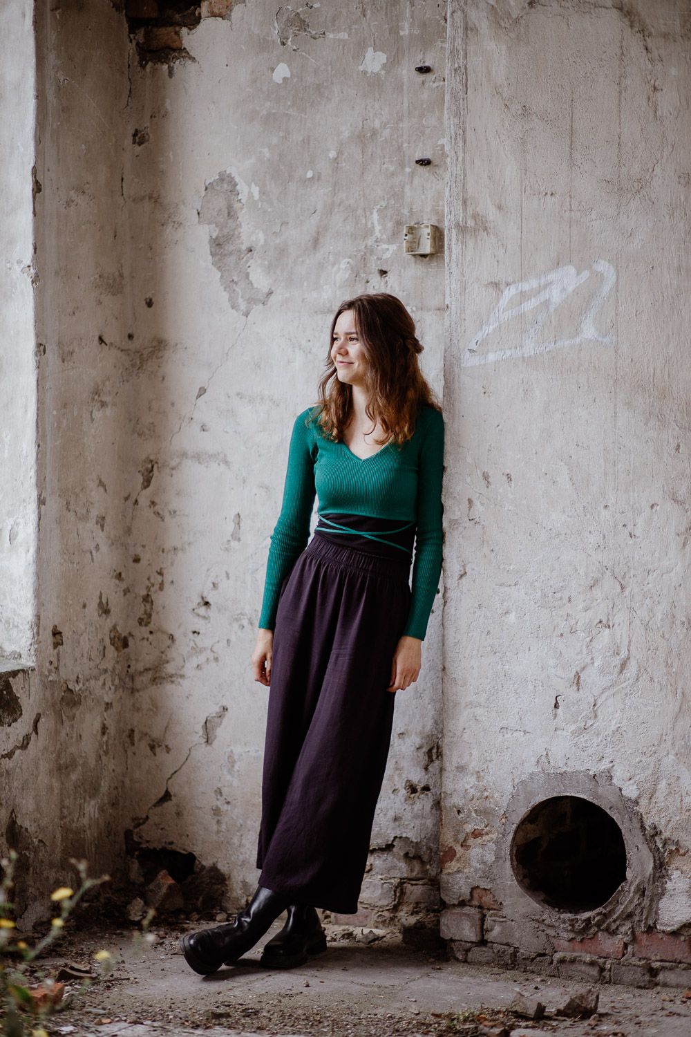 Young woman in green sweater and purple wide-leg pants posing in abandoned building.