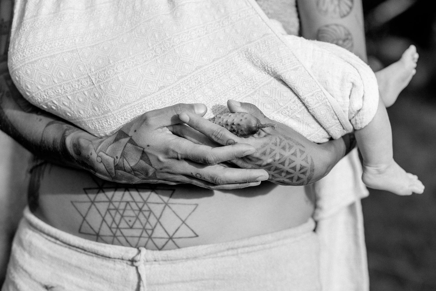 Black and white close-up of tattooed hands holding newborn baby.