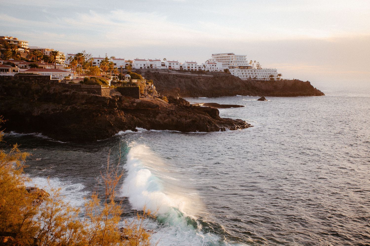Dramatic coastal sunset view of white buildings perched on volcanic rocky cliffs above crashing ocean waves.