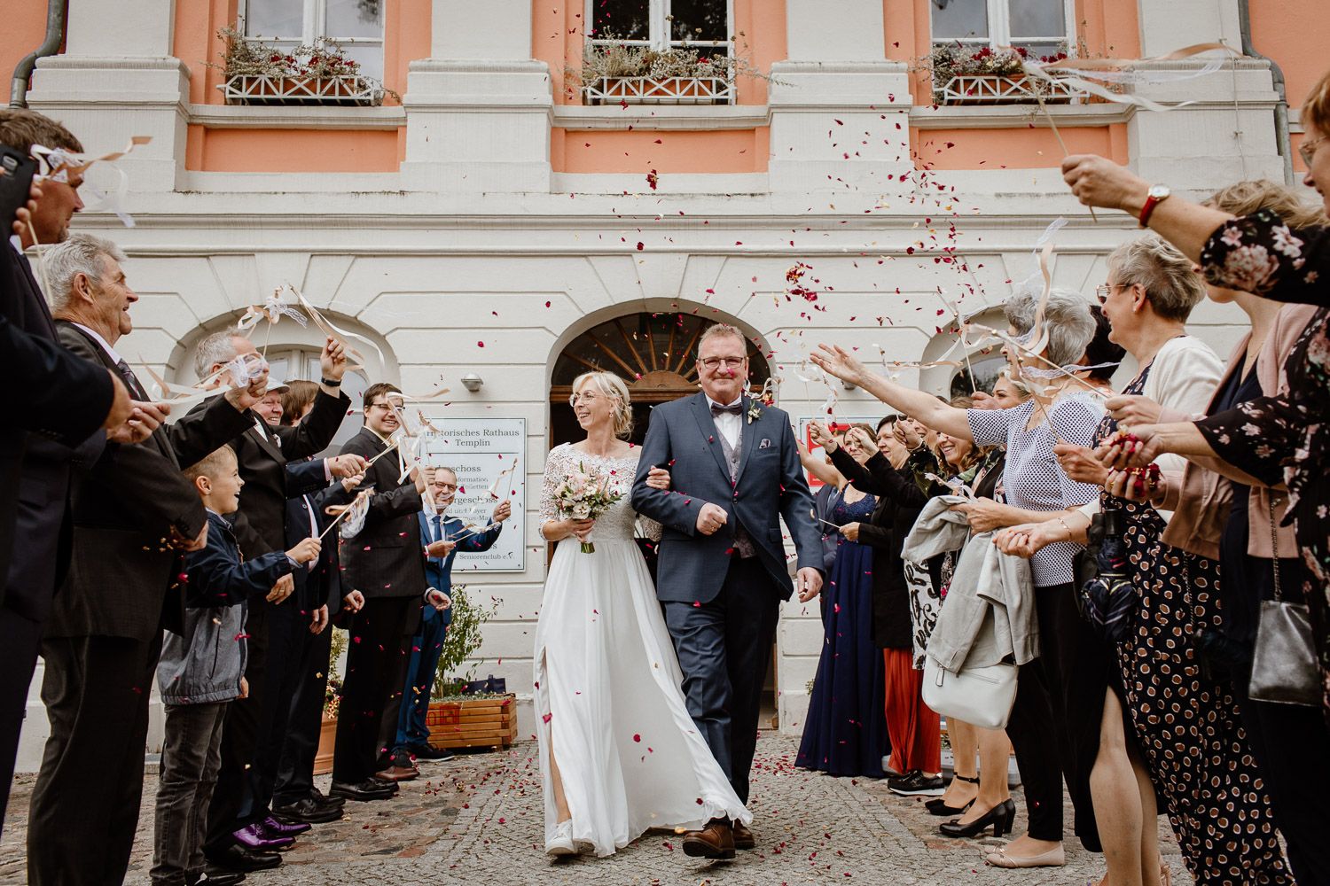 Newlyweds walk through confetti shower thrown by guests outside elegant historic building.