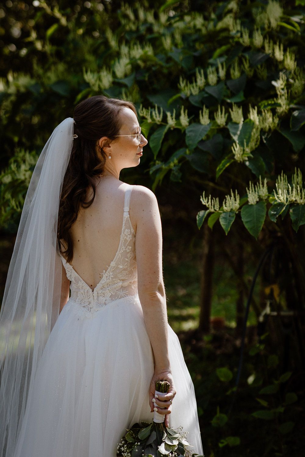 Bride in backless lace wedding gown with veil holding bouquet, standing before lush green foliage.