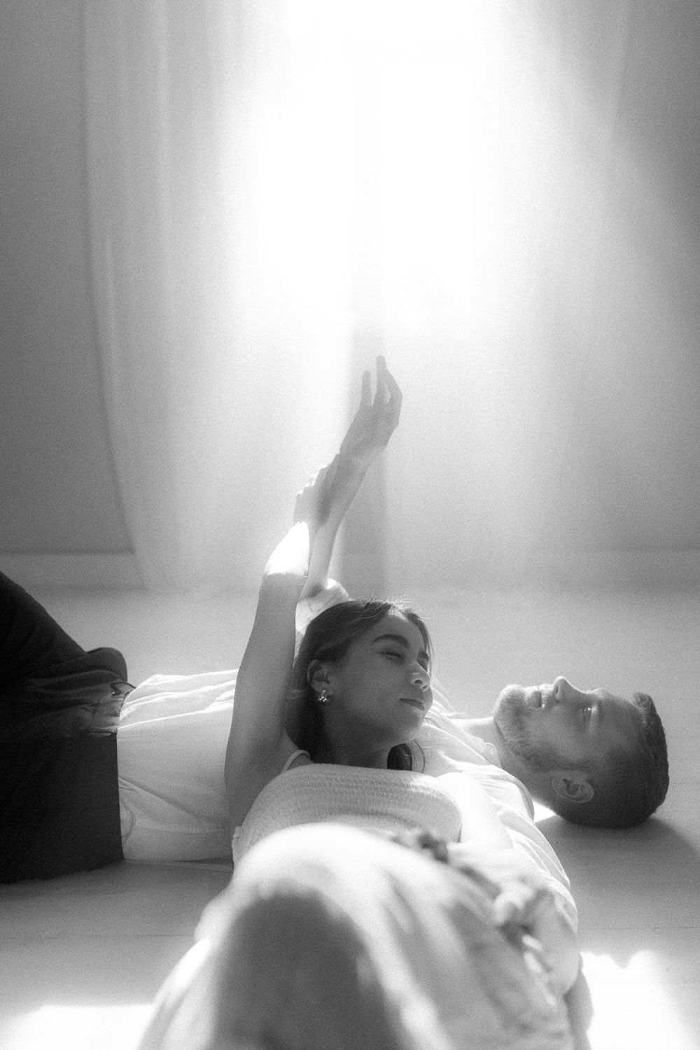 Black and white artistic photo of a couple lying on the floor bathed in dramatic bright light.