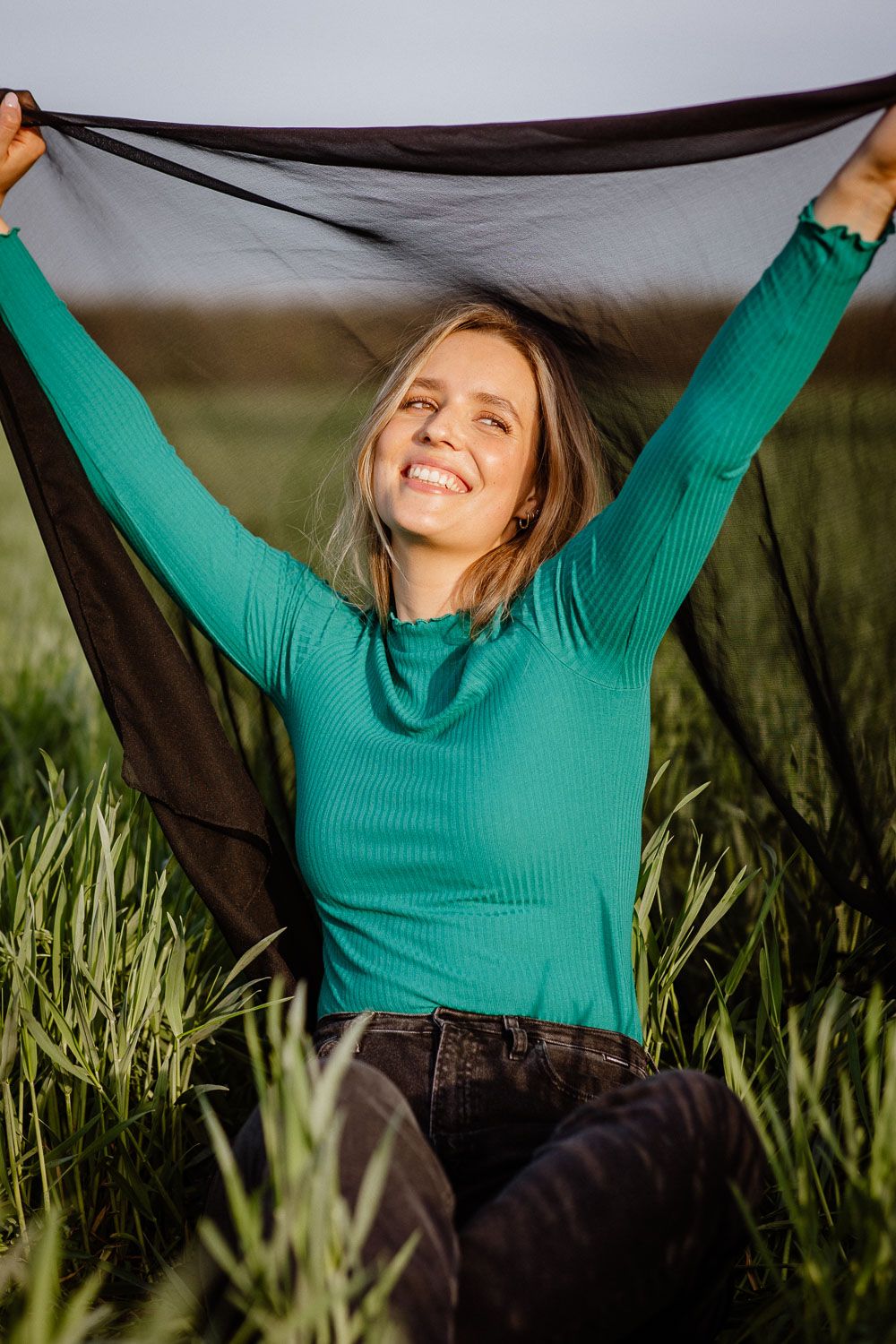 Happy blonde woman in teal top holding black fabric overhead while sitting in green grass.