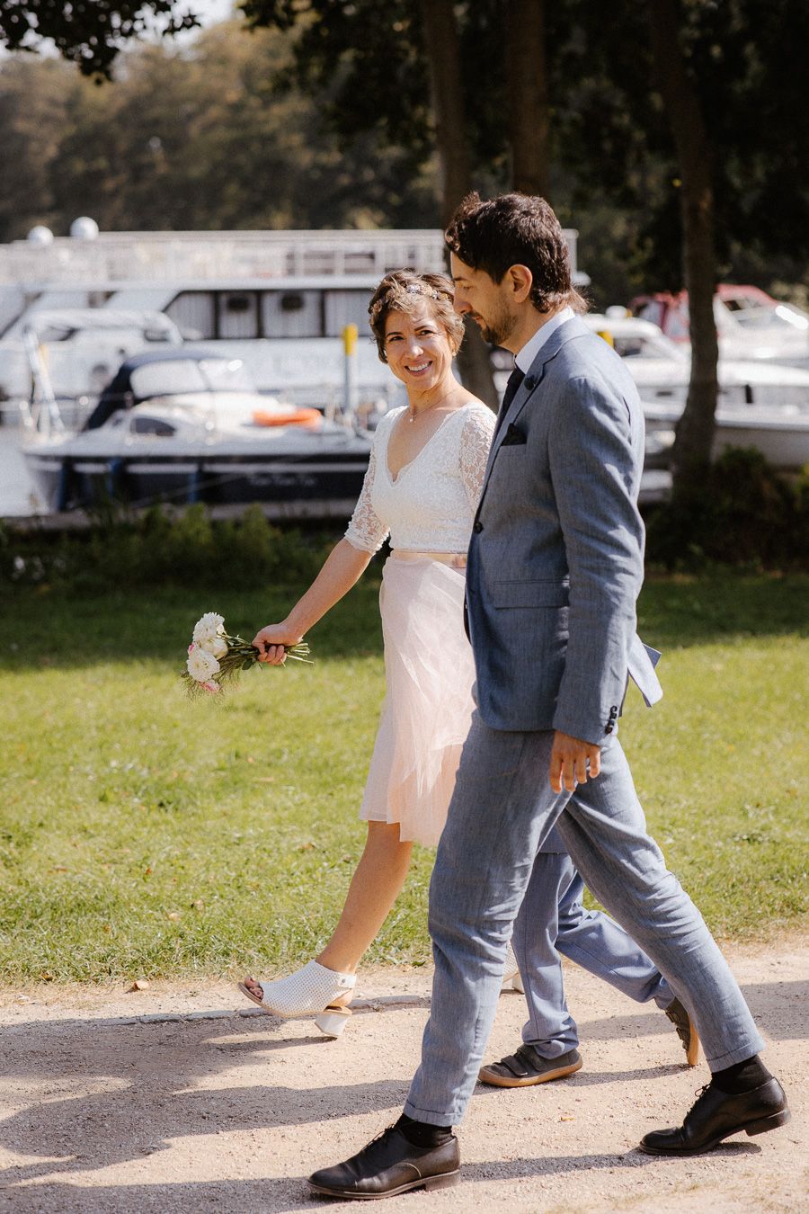 Smiling bride in short white dress walks with groom in blue suit near marina on sunny day.