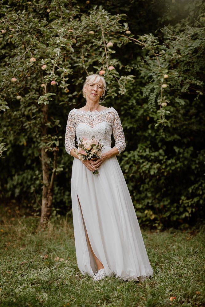 Mature bride in lace and chiffon wedding gown holding bouquet, standing before an apple tree.