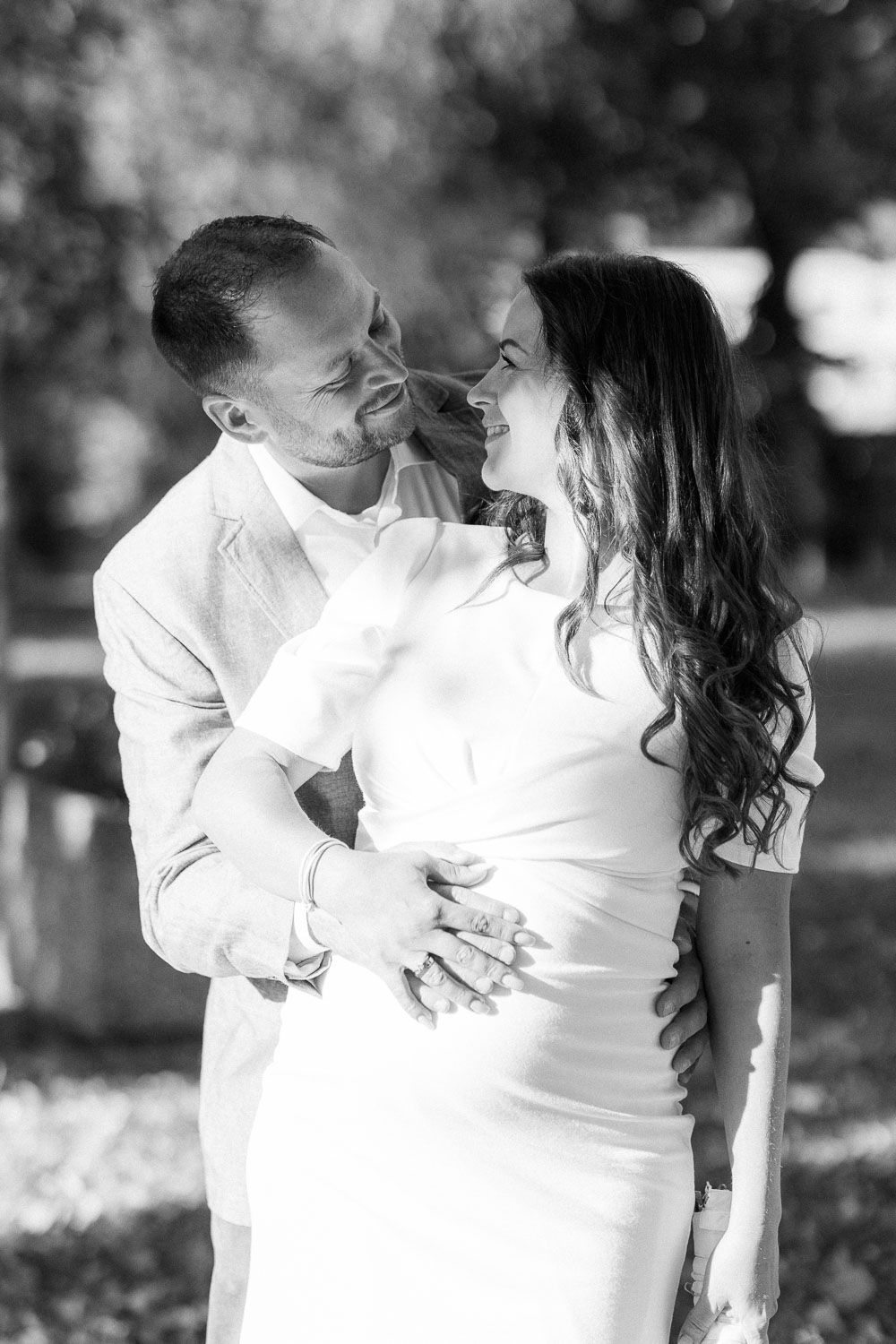 Black and white portrait of a smiling couple embracing tenderly in a sunlit outdoor park setting.