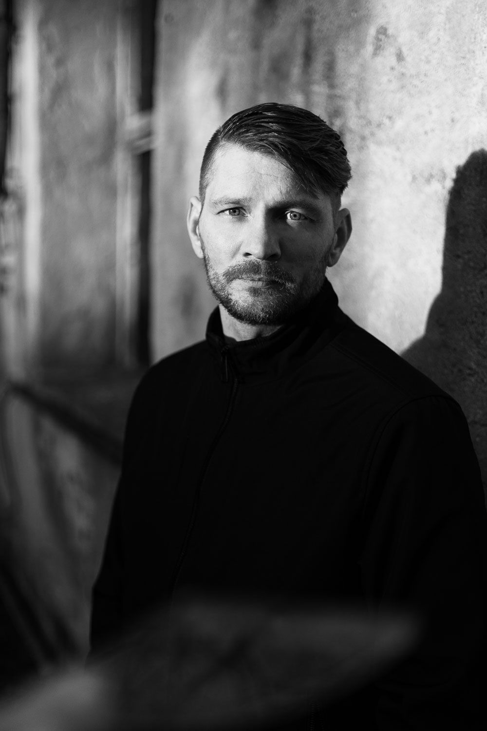 Black and white portrait of a bearded man in a black turtleneck leaning against a textured wall.
