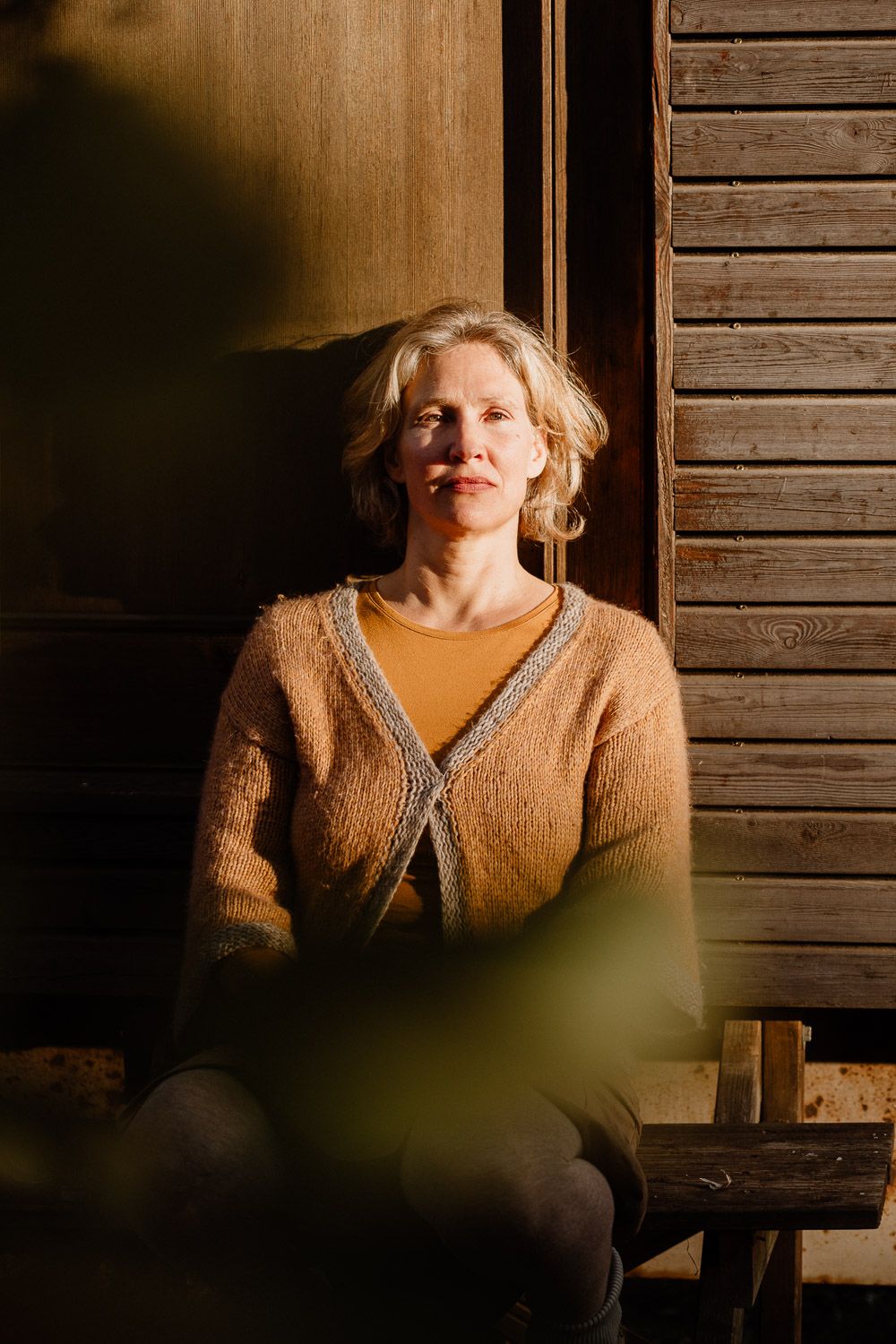 Blonde woman in cozy orange sweater sitting in warm sunlight by wooden wall.
