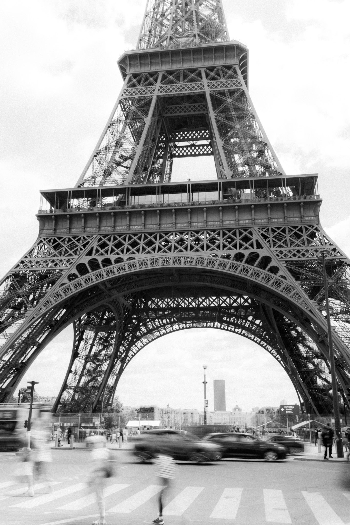 Black and white photo of the Eiffel Tower base with blurred traffic and pedestrians in Paris, France.