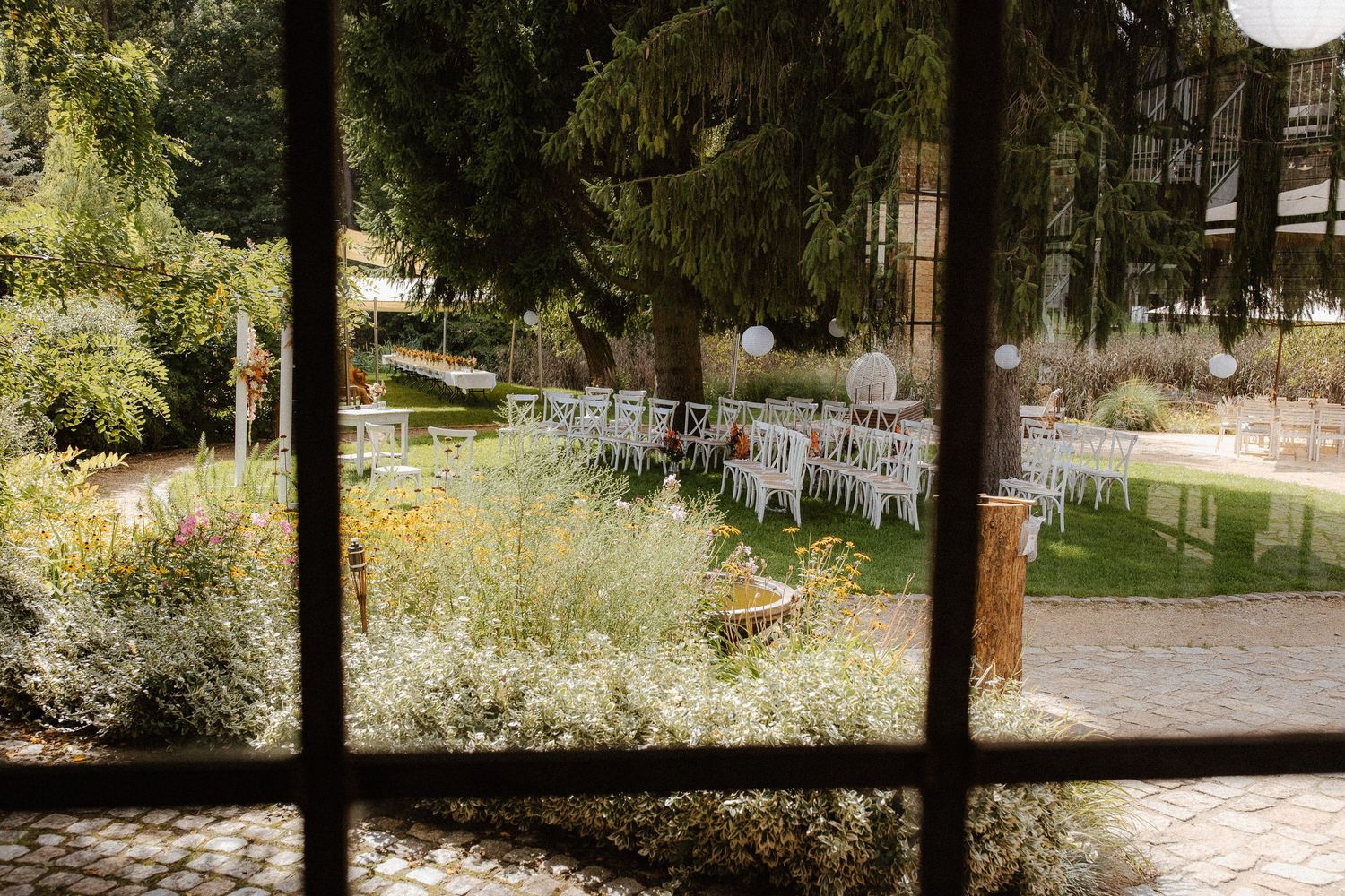 Outdoor wedding ceremony setup with white chairs arranged in rows amid lush garden greenery and floral decorations.