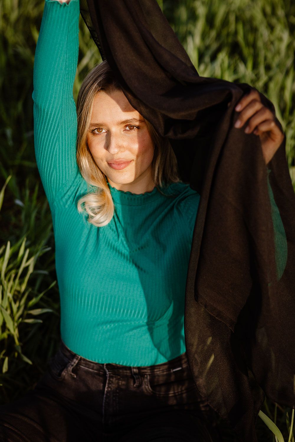 Young blonde woman in teal ribbed top holding black fabric aloft in a sunny green field.