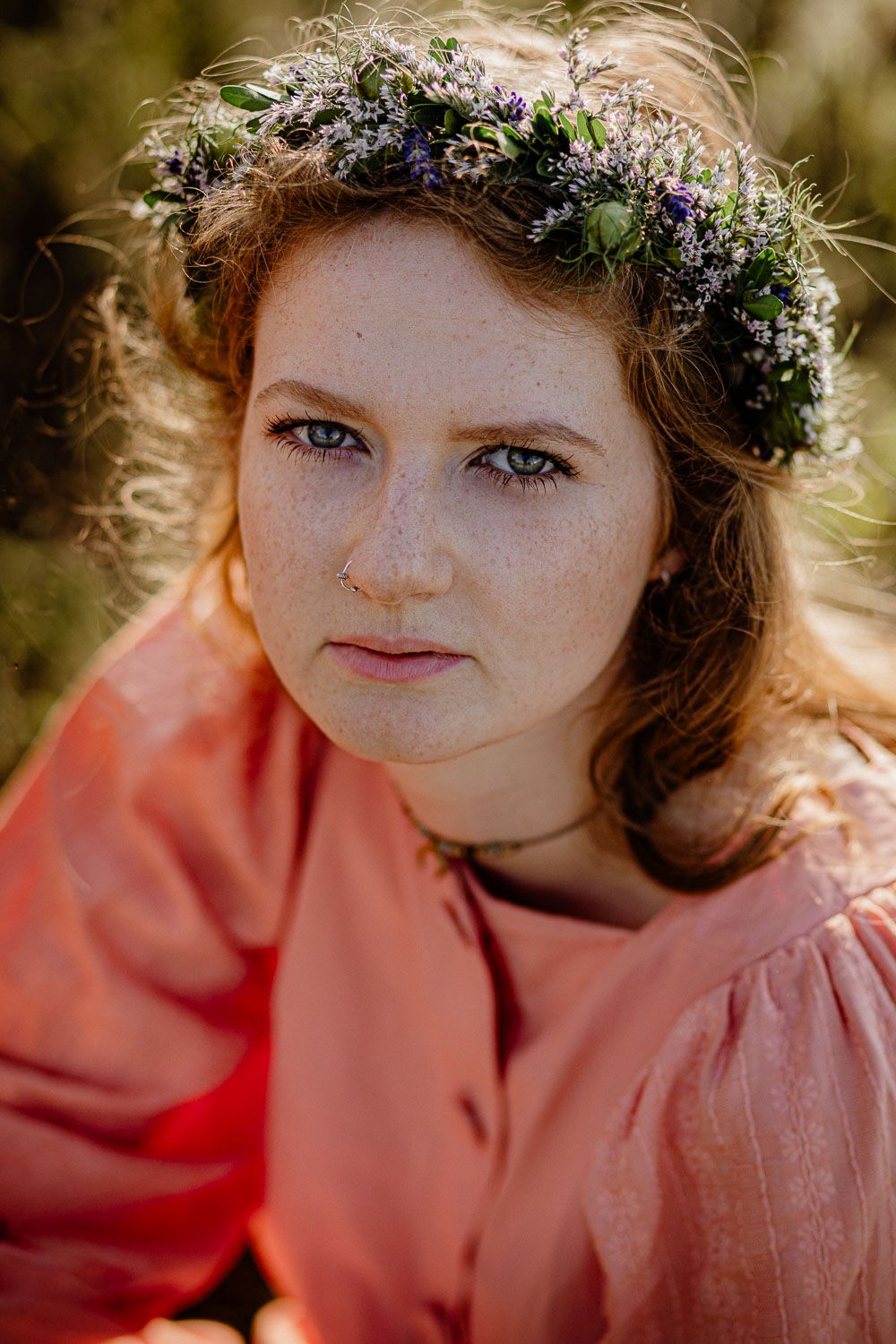 Close-up portrait of a young woman with freckles wearing a floral crown and pink dress outdoors.