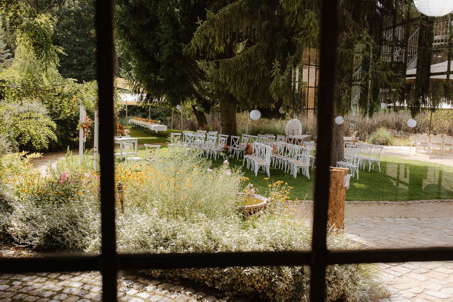 Outdoor wedding ceremony setup with white chairs arranged in garden viewed through rustic window frame.