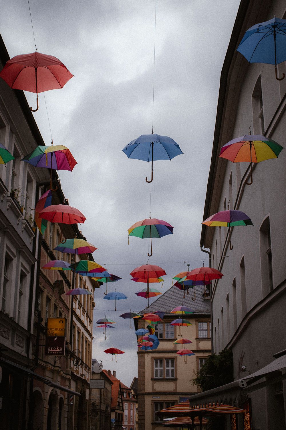 Colorful rainbow umbrellas suspended between historic European buildings under a dramatic overcast sky.