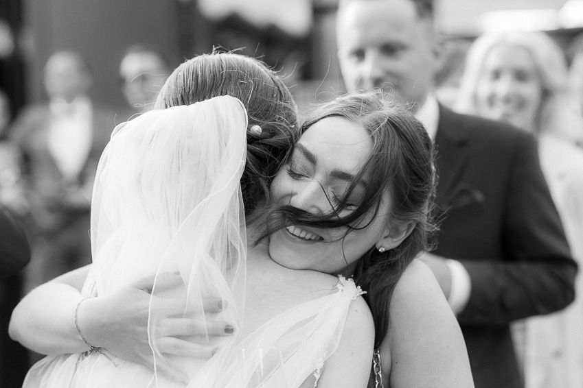 Bride and bridesmaid share a warm emotional hug during wedding celebration in black and white photo.