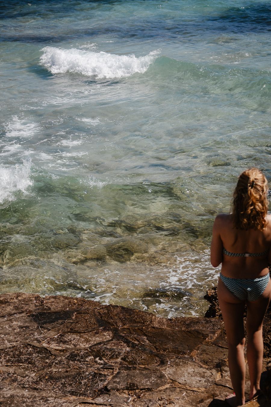 Woman in bikini standing on rocky shoreline overlooking clear turquoise ocean waves.
