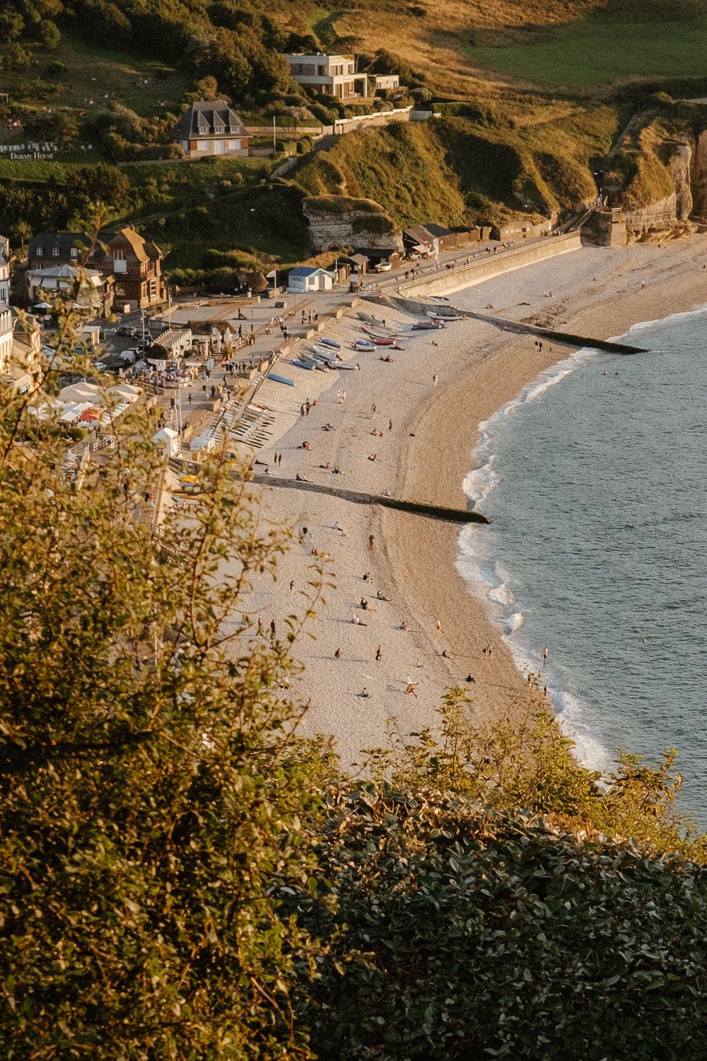 Aerial golden hour view of a coastal village beach with cliffs, houses, and crowds in warm sunlight.
