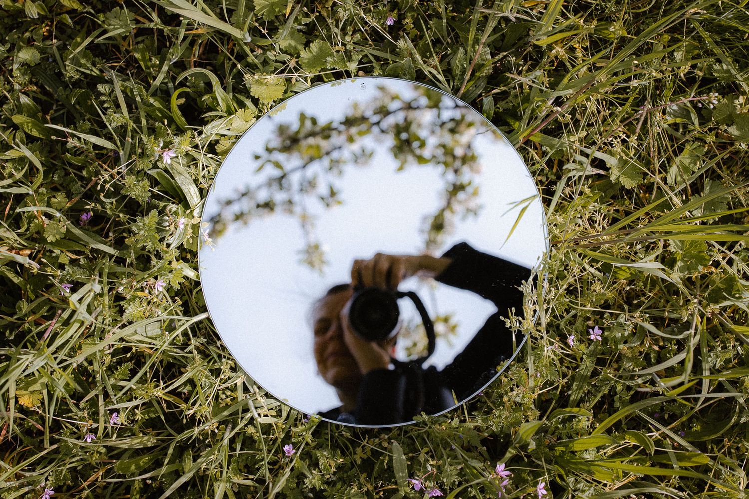 Round mirror lying in green grass reflecting photographer taking self-portrait with camera overhead.