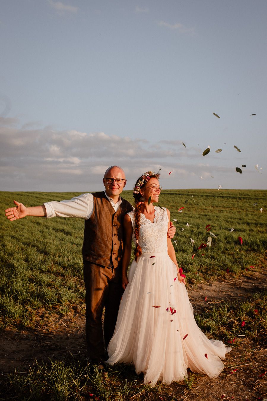 Joyful wedding couple celebrating outdoors in a green field with rose petals falling around them at golden hour.