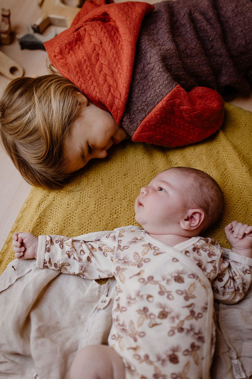 Toddler gently nuzzling a newborn baby lying on a yellow blanket indoors.