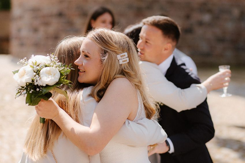 Bride in white gown holding bouquet sharing a warm embrace with a guest while groom hugs another nearby.