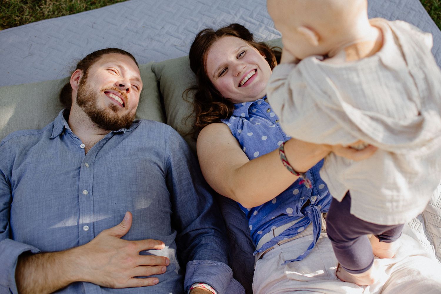 Joyful family of three lying on blanket outdoors with baby standing.