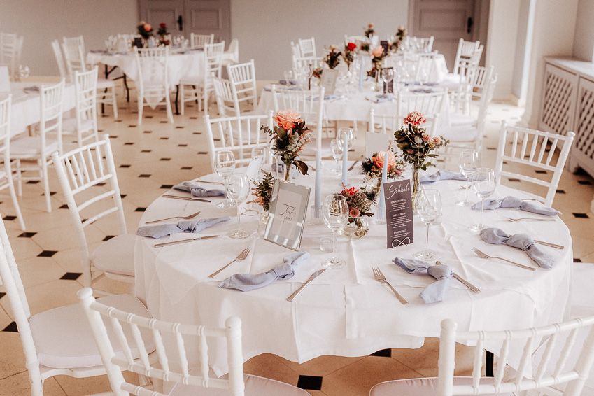 Elegant wedding reception venue with round tables, white linens, chiavari chairs, and floral centerpieces on a checkered floor.