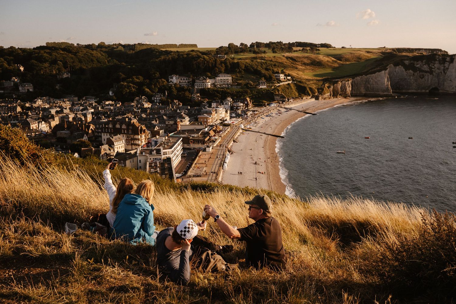 Group of friends sitting on grassy cliff overlooking charming coastal village beach at sunset.