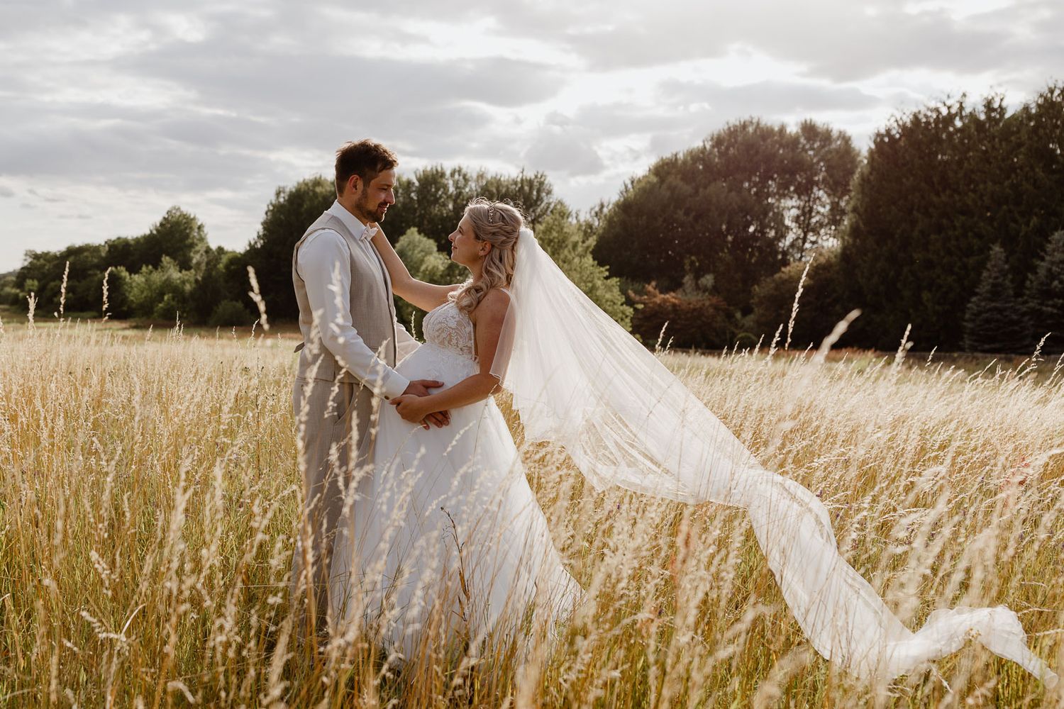 Bride and groom embrace romantically in golden wheat field with veil flowing in wind.