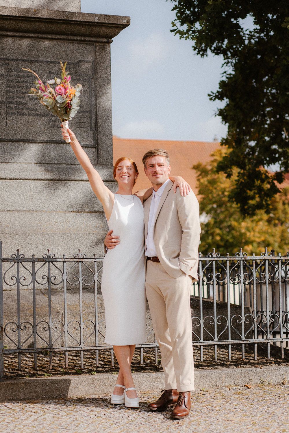 Joyful bride in white midi dress raises colorful bouquet while groom in beige suit smiles outdoors.