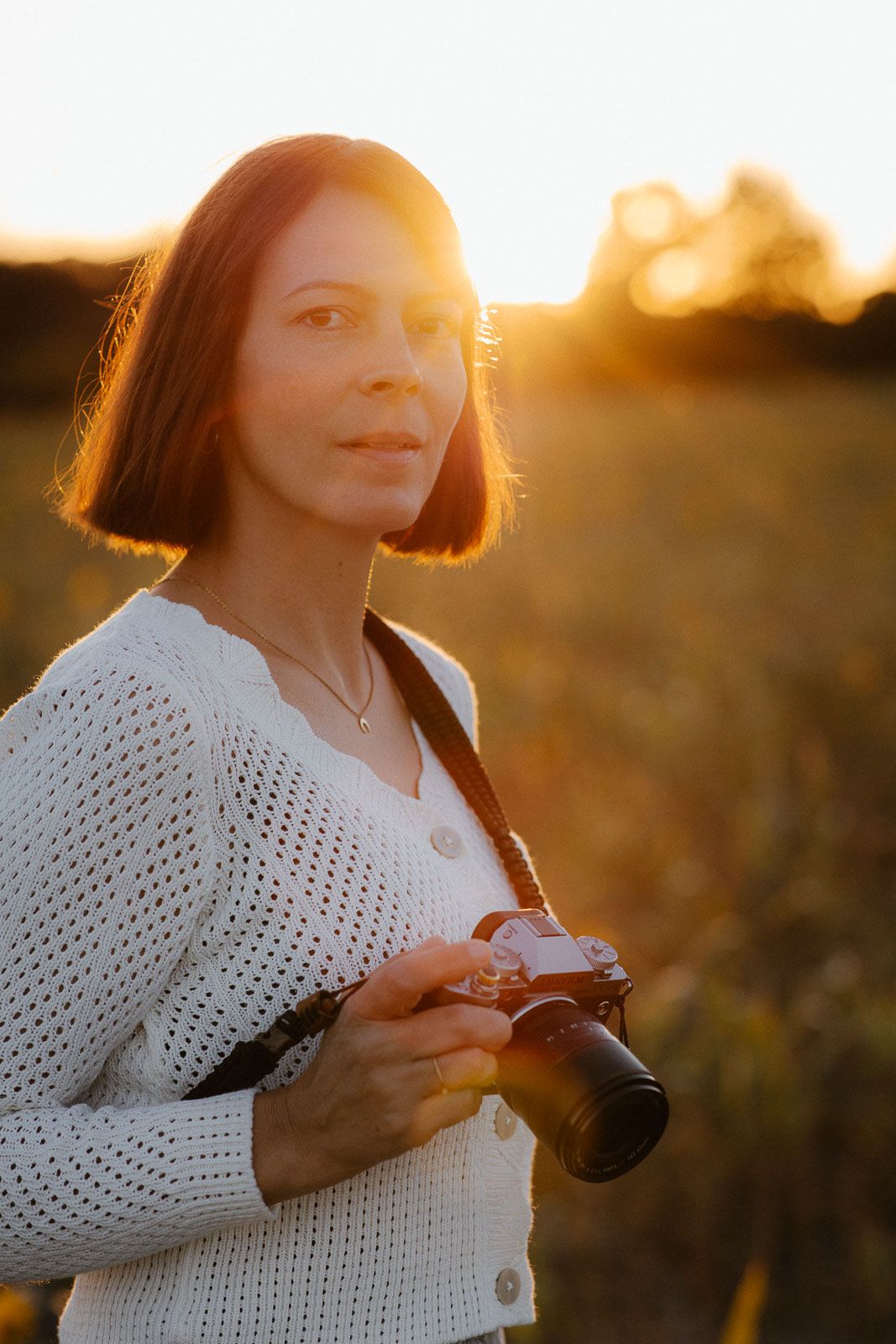 Portrait of a woman holding a film camera backlit by golden sunset light in an open field.