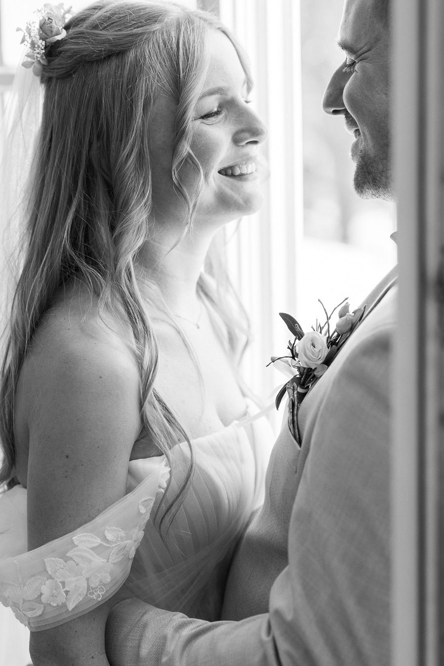 Joyful bride in off-shoulder wedding dress laughs with groom in black and white romantic portrait.
