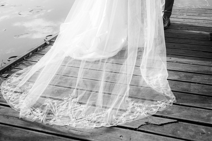 Elegant lace-trimmed wedding dress train spreads across rustic wooden dock in black and white photo.
