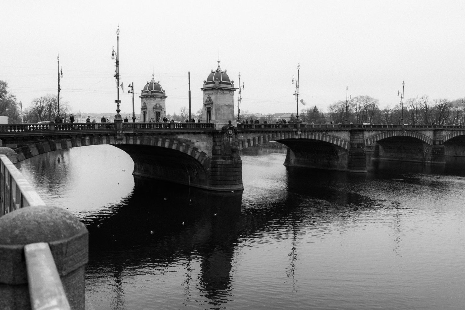 Black and white photo of a historic stone bridge with arched spans reflecting in calm river water on overcast day.