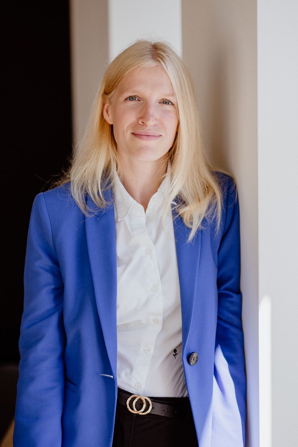 Professional blonde woman in blue blazer and white shirt leaning against a wall smiling.