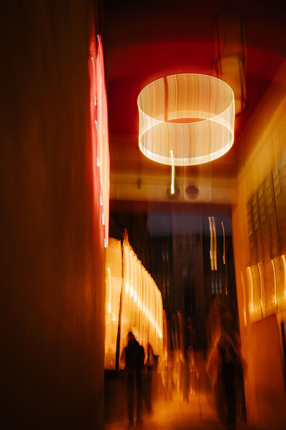 Long exposure shot of glowing cylindrical lamp in warmly lit corridor with blurred silhouettes.