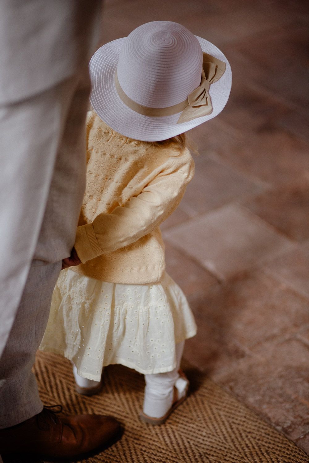 Young flower girl in yellow dress and white hat standing on stone floor at a wedding.