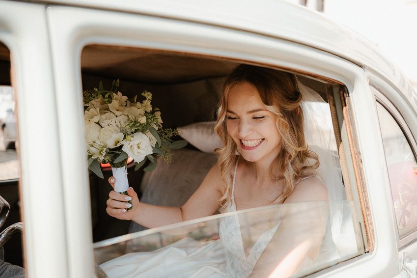 Smiling bride in white gown holding a white floral bouquet while sitting inside a vintage cream-colored car.