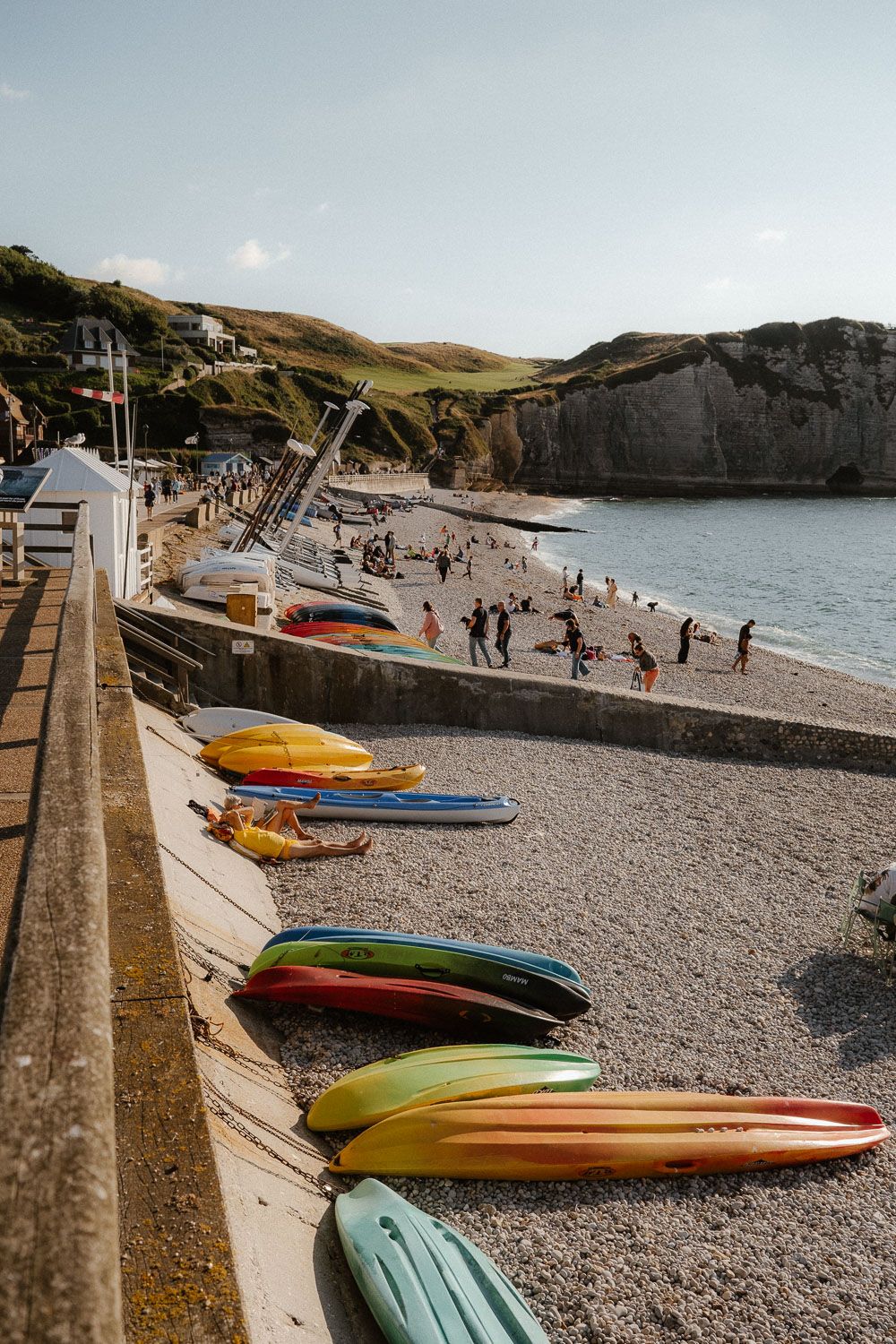 Colorful kayaks lined up on pebble beach with swimmers and white chalk cliffs in background.