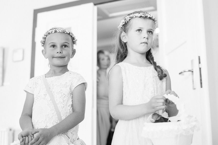 Two young flower girls in white dresses and floral crowns standing at a doorway during a wedding ceremony.