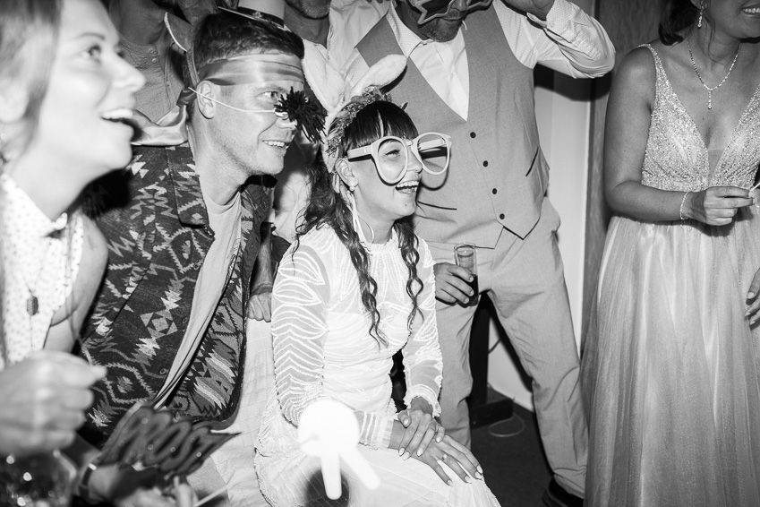 Bride and groom wearing decorative masks laughing with guests during a festive wedding party celebration.