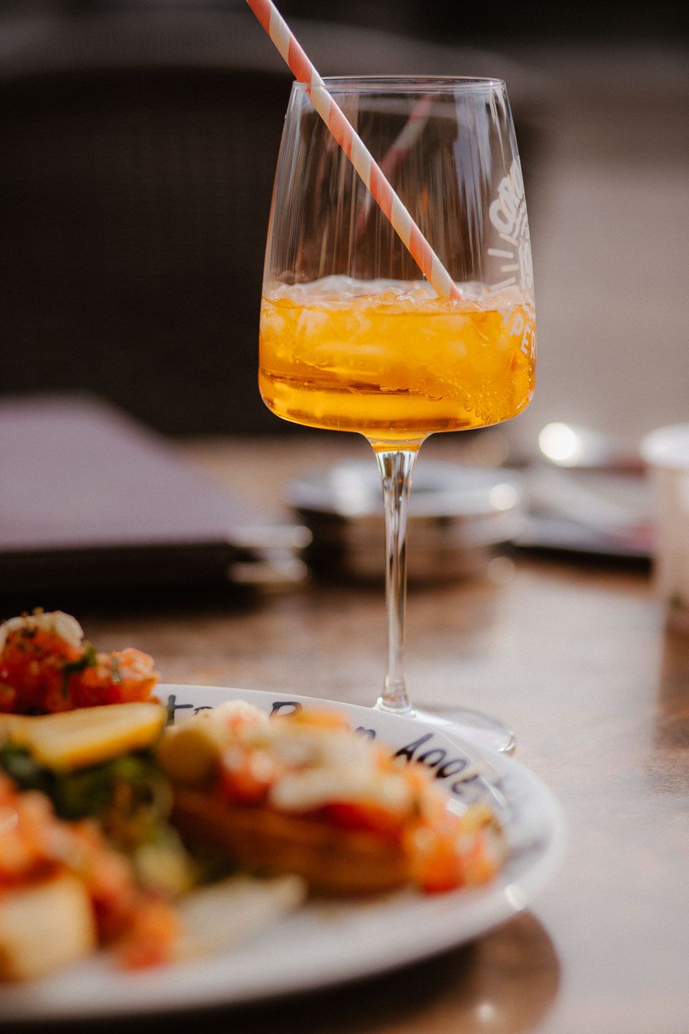 A glass of orange Aperol Spritz cocktail with a straw sits beside a plate of bruschetta on a restaurant table.