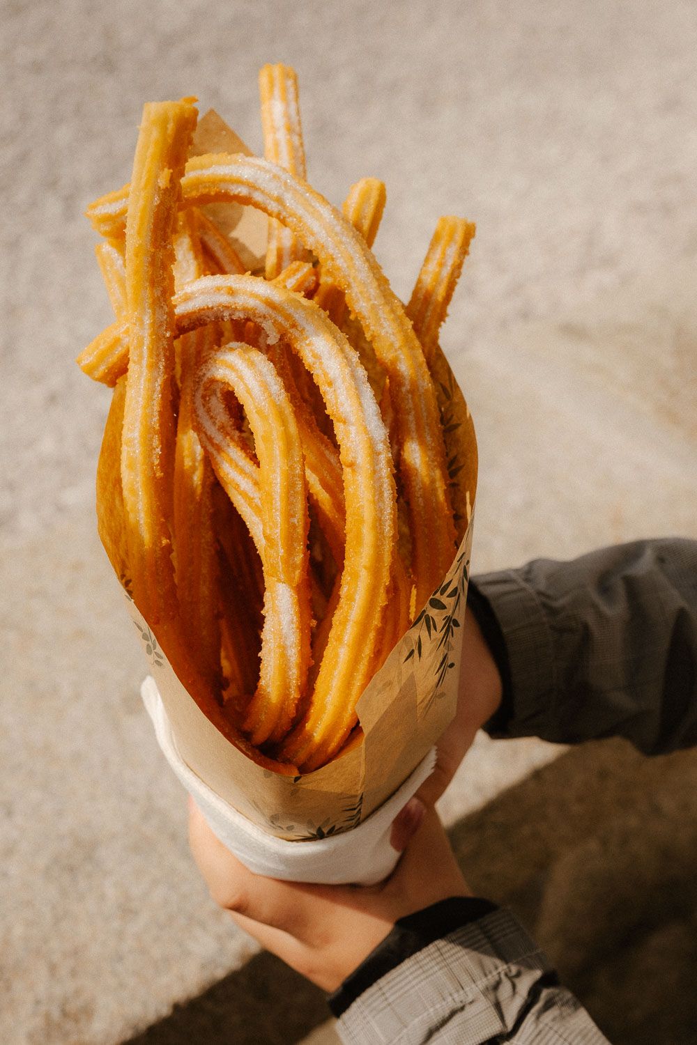 Hands holding a paper cup filled with fresh churros dusted with powdered sugar outdoors.